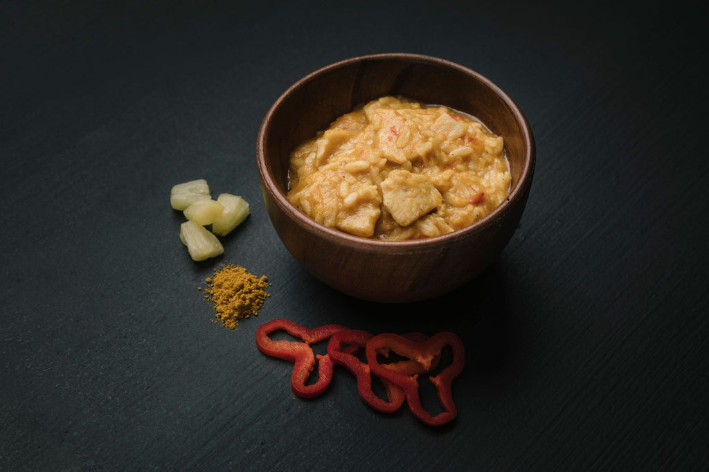 Bowl of Real Turmat Chicken Curry on a dark surface with spices and red pepper slices.