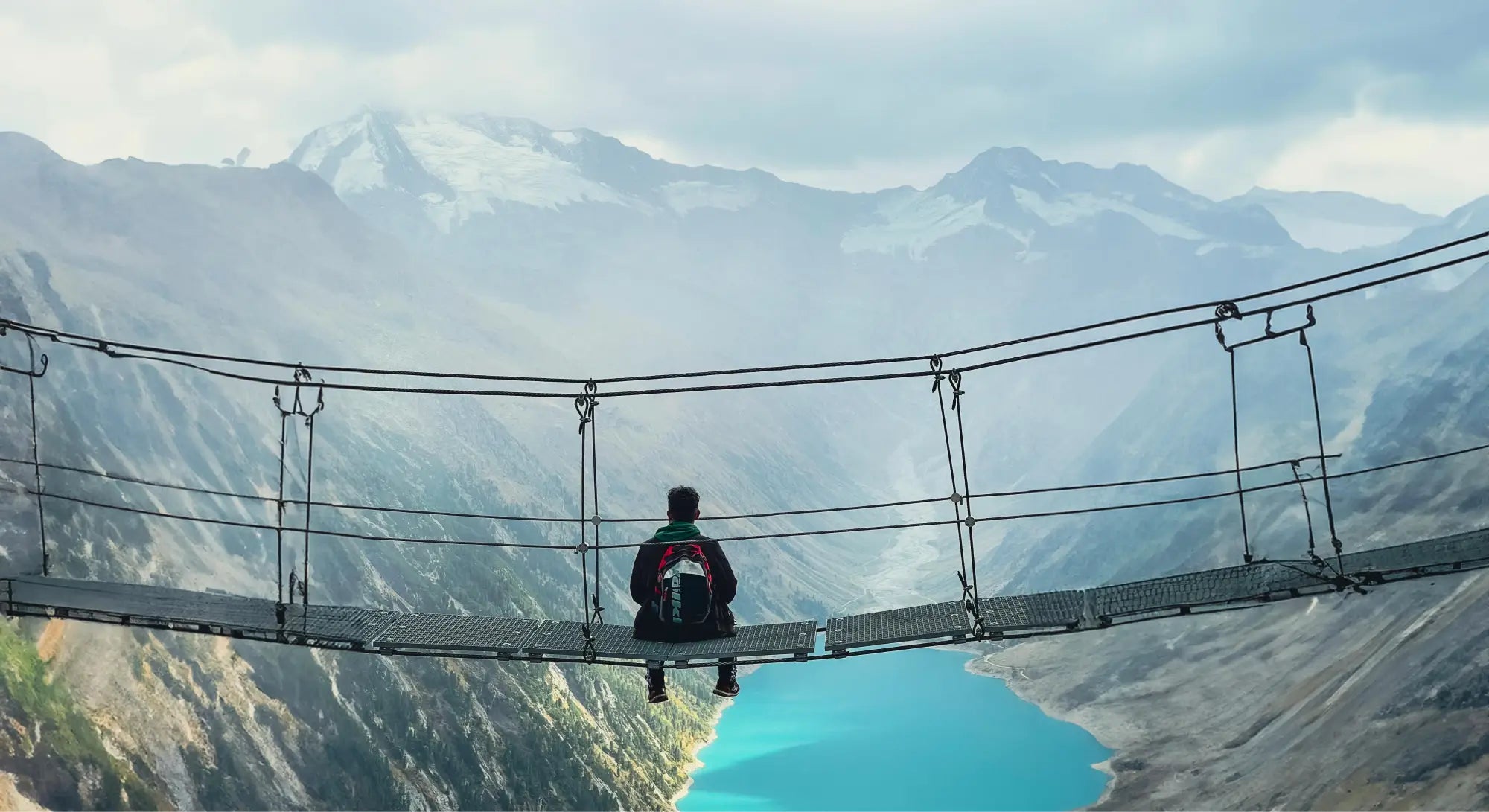 Person sitting on a suspension bridge overlooking a mountainous landscape with a lake.