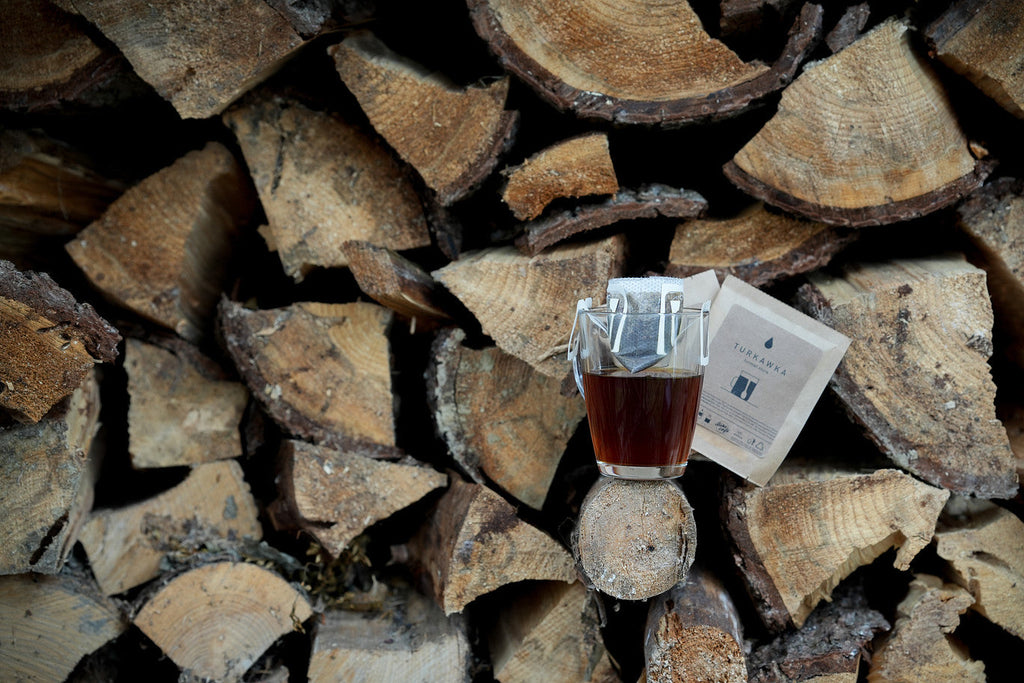 Two packets of drip bag coffe Turkawka with a small cup of coffe on a background of stacked wooden logs.