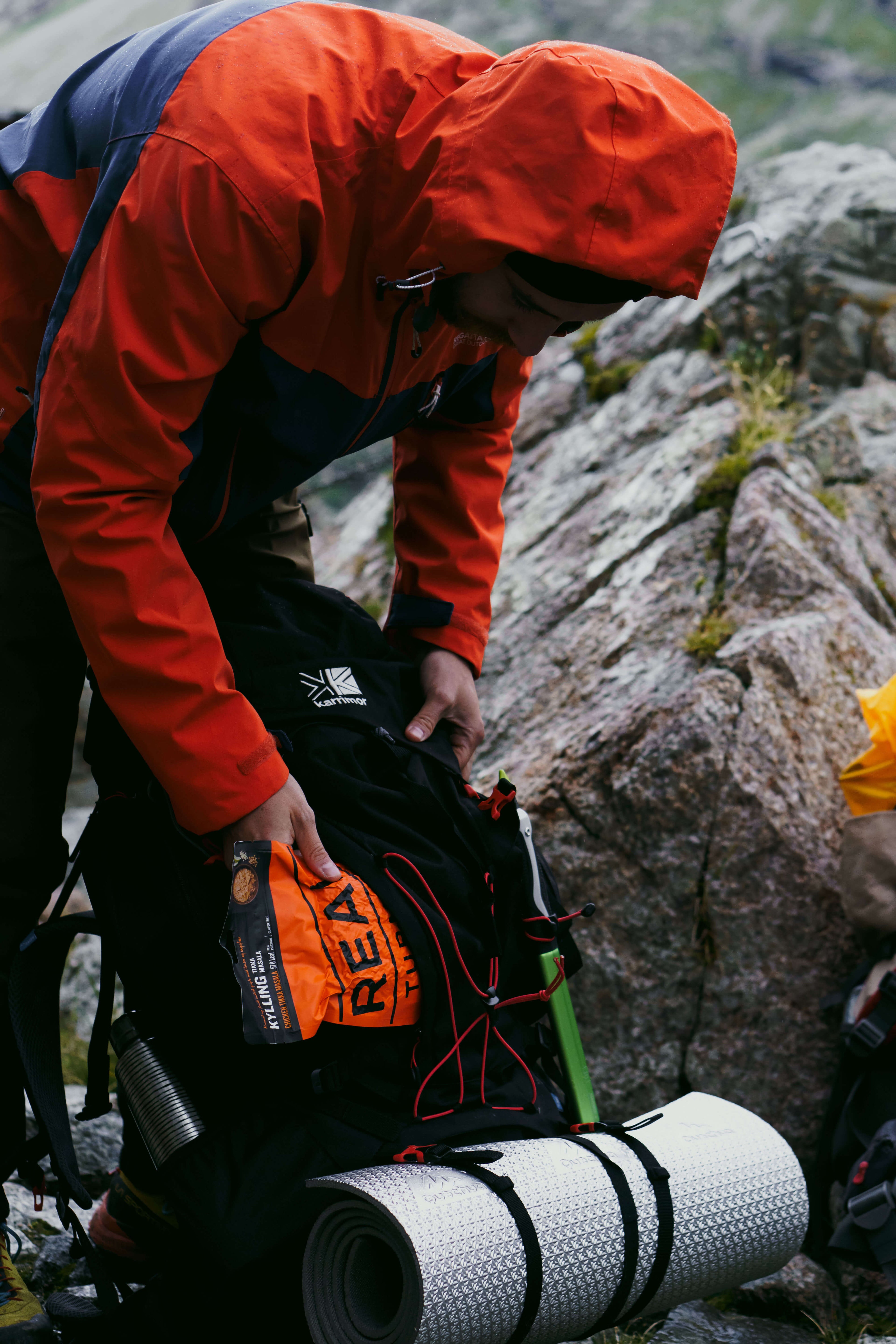 Person in orange jacket with backpack and camping gear on rocky terrain with Real Turmat Chicken Tikka Masala