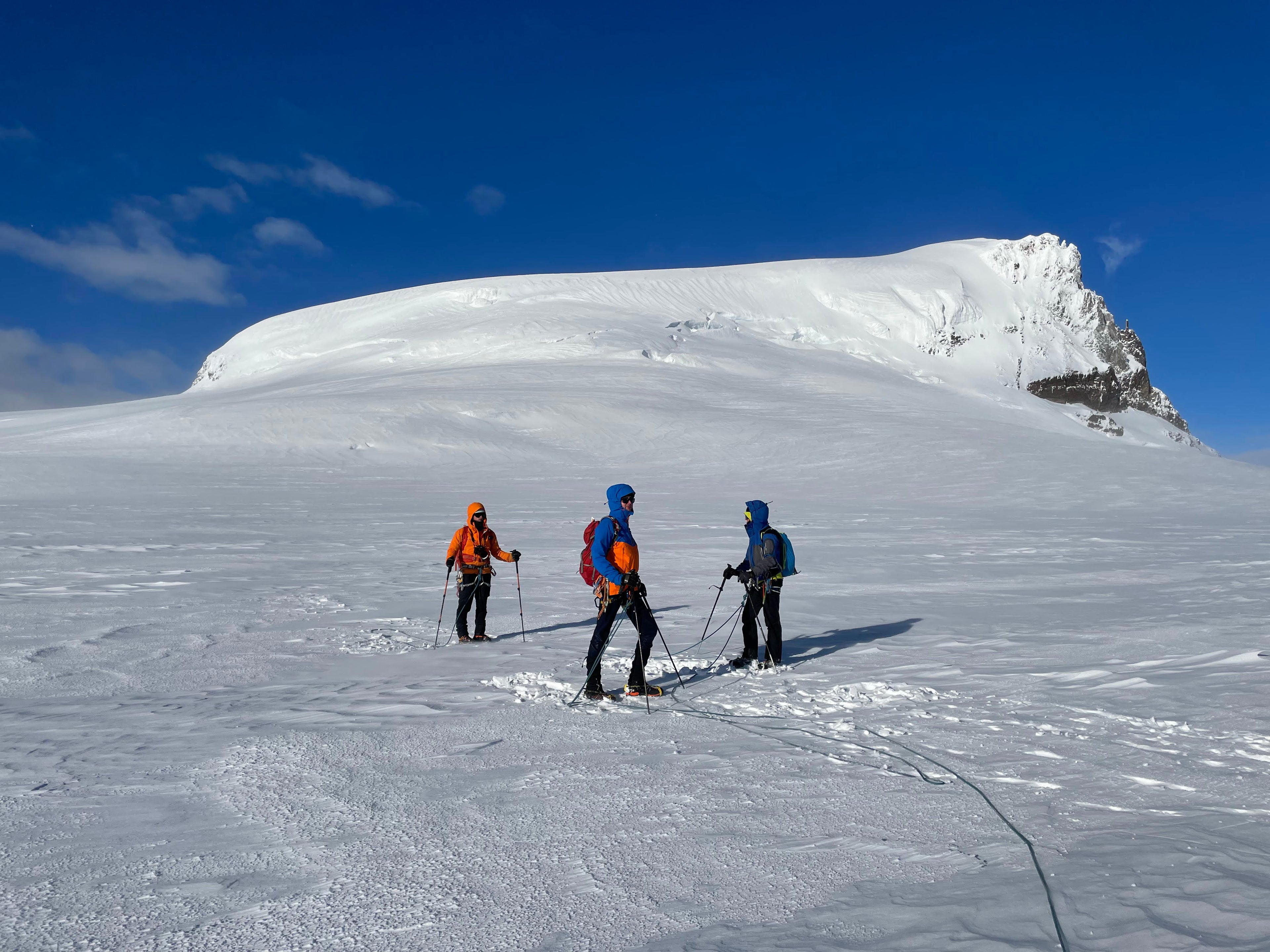 Three mountaineers with poles and backpacks trekking across a snowy mountain slope under a clear blue sky, with a snow-covered peak in the background.