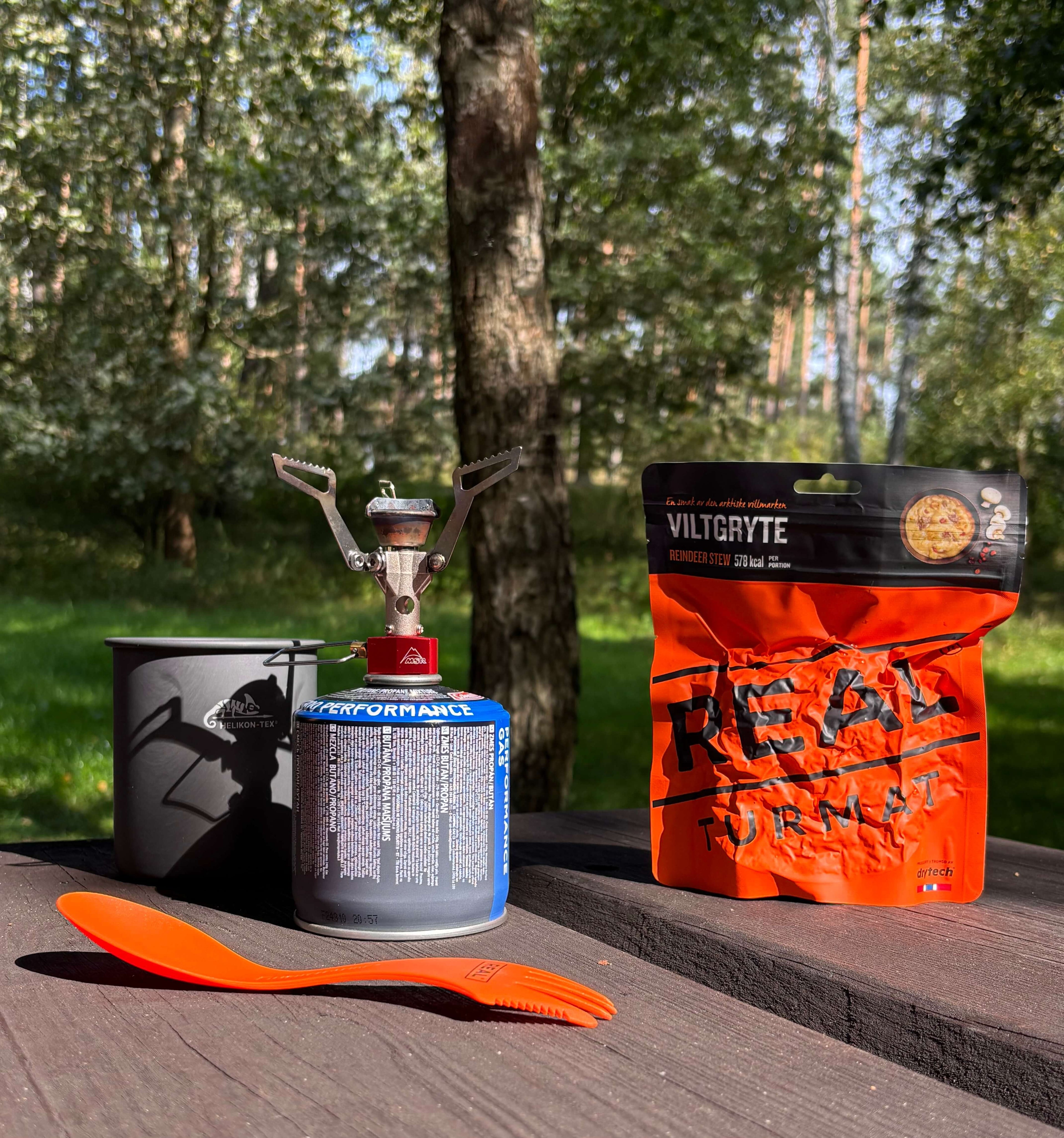 Camping stove, mug, and Real Turmat Reindeer Stew meal package on a wooden surface with a forest background