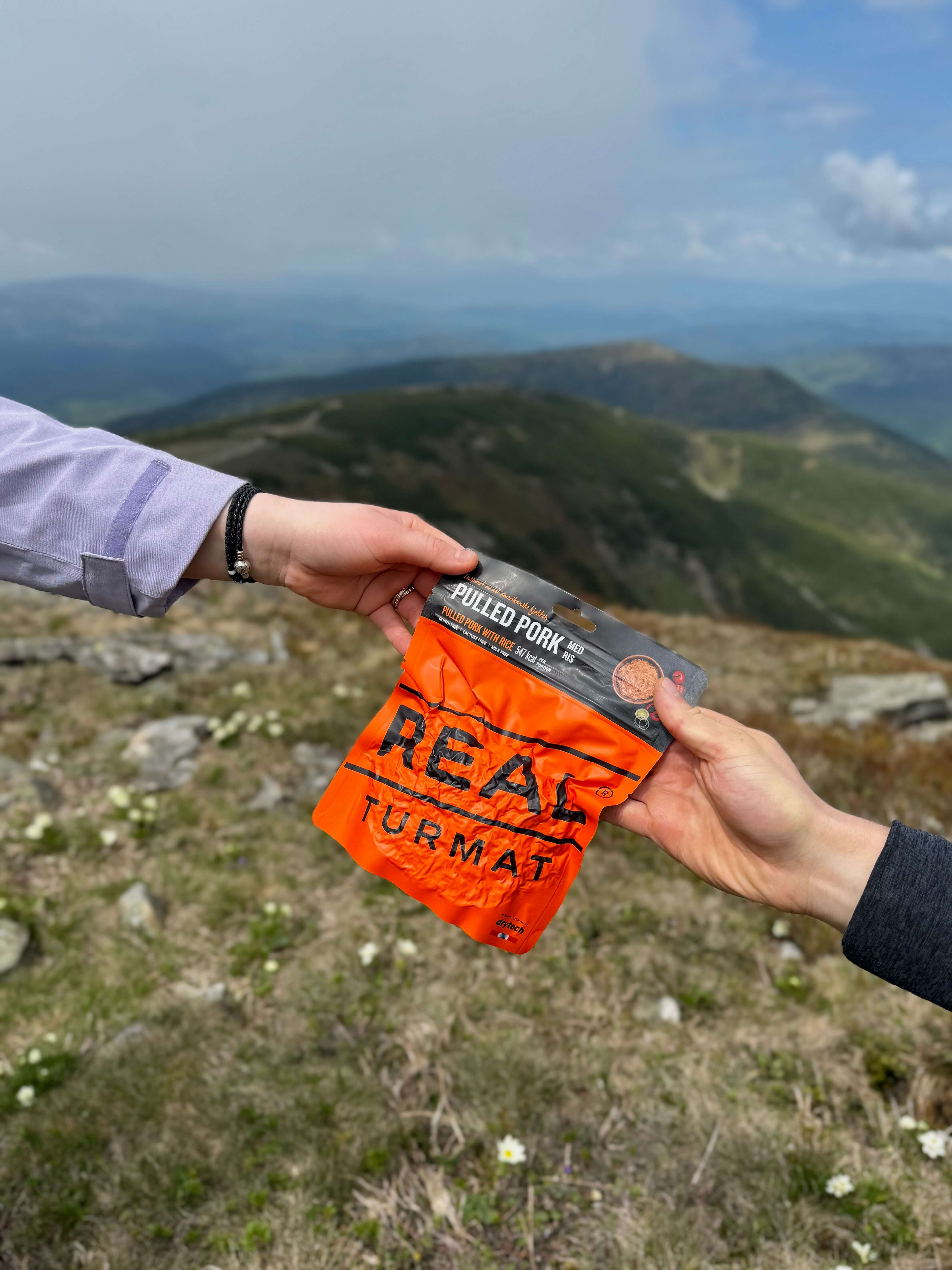 Two hands exchanging a package of 'Real Turmat' pulled pork over a scenic mountain landscape.