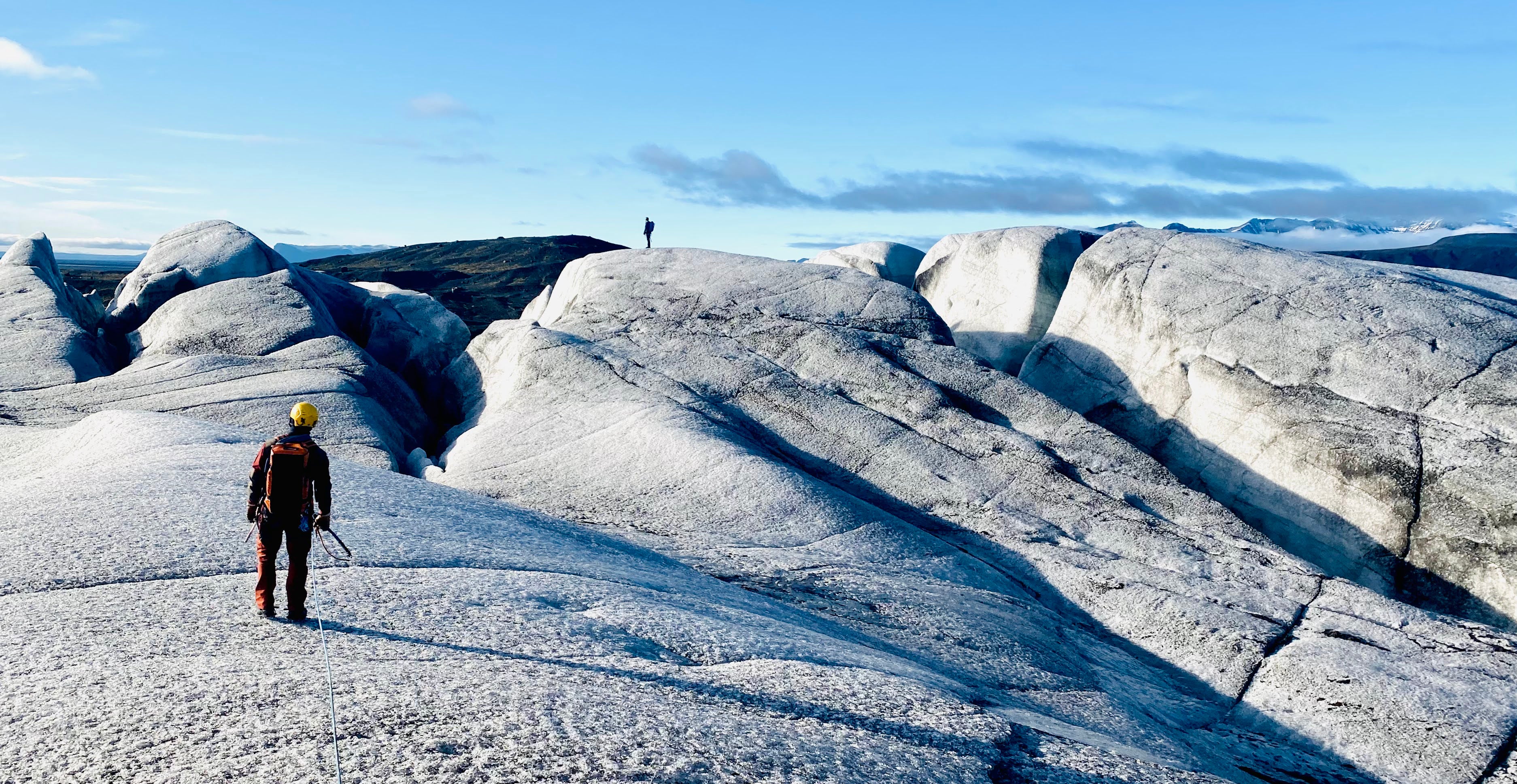 A person in outdoor gear stands on a snowy glacier landscape with large ice formations under a blue sky, with another distant figure visible in the background.