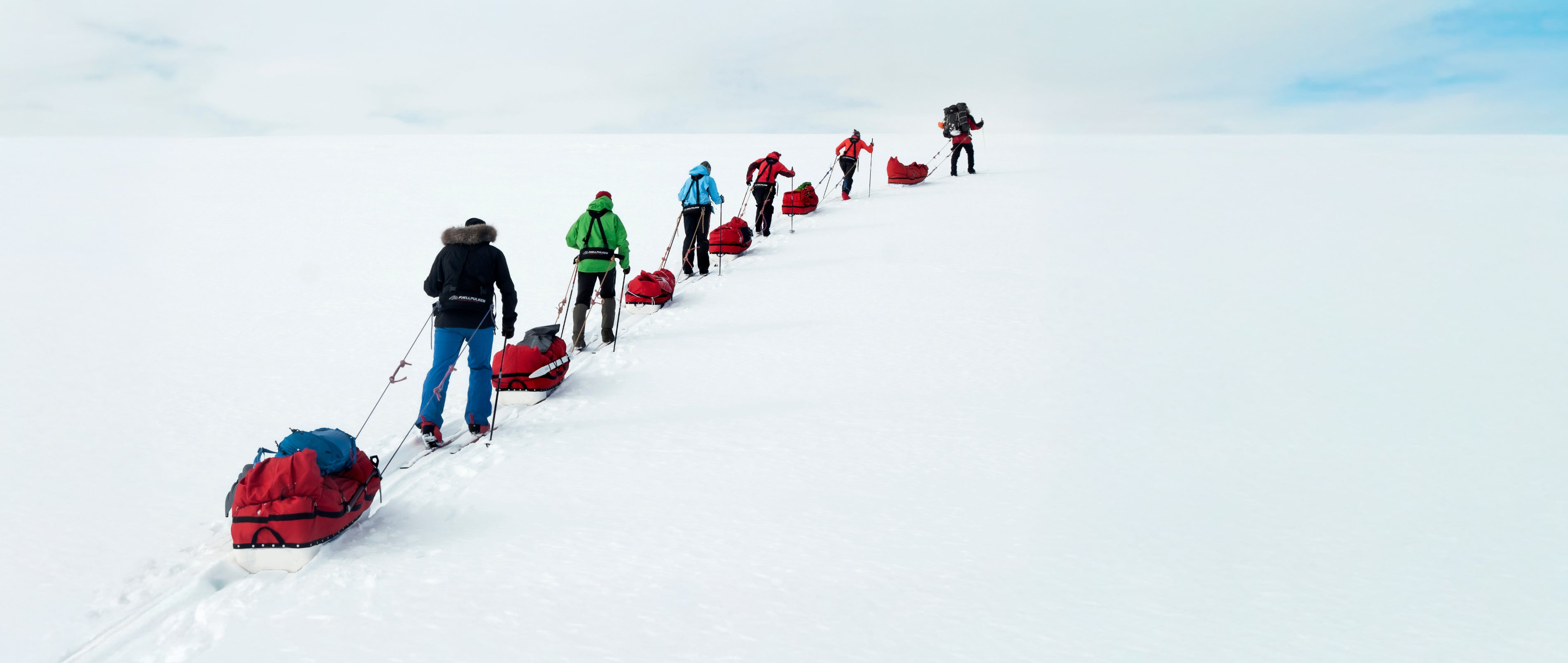 A long line of people with sleds and red backpacks traveling across a vast, snow-covered landscape under a pale sky.