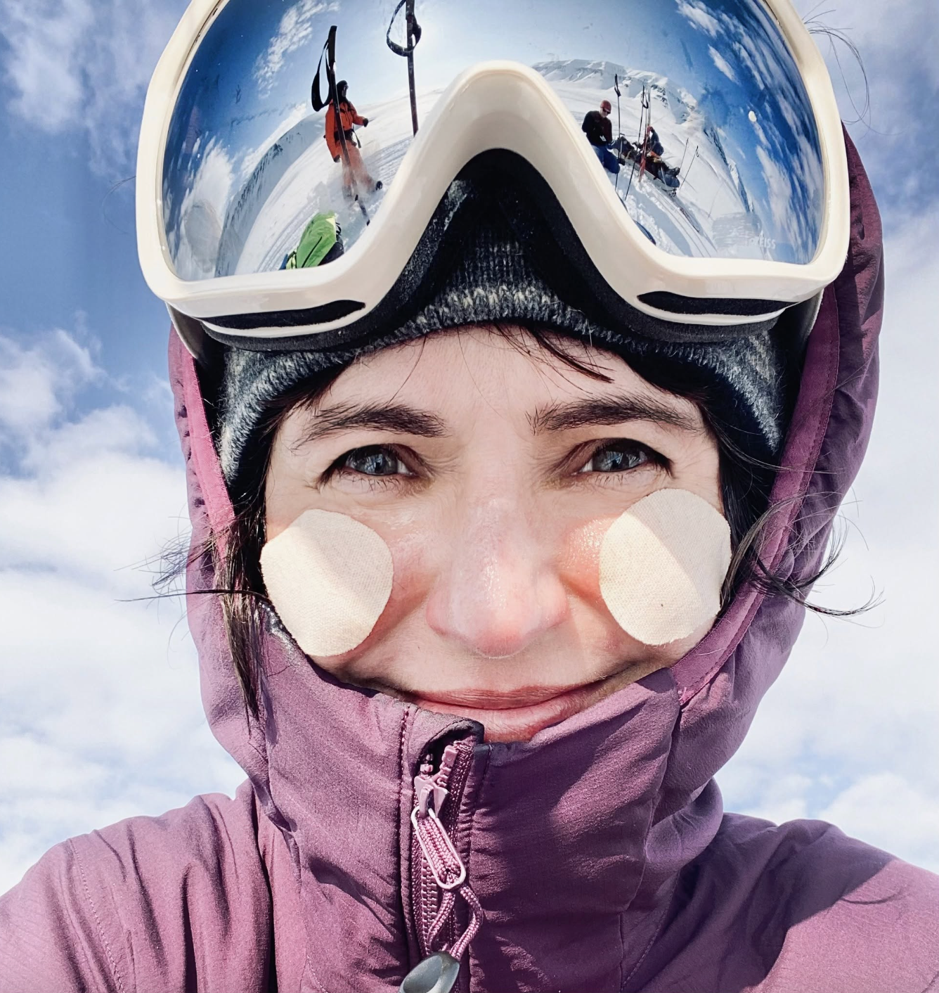 Anita Horowska
Traveler and photographer - A close-up selfie of a person wearing ski goggles with a mirrored sky reflection, a black beanie, and a purple hooded jacket. Two round beige patches are applied to the cheeks. 